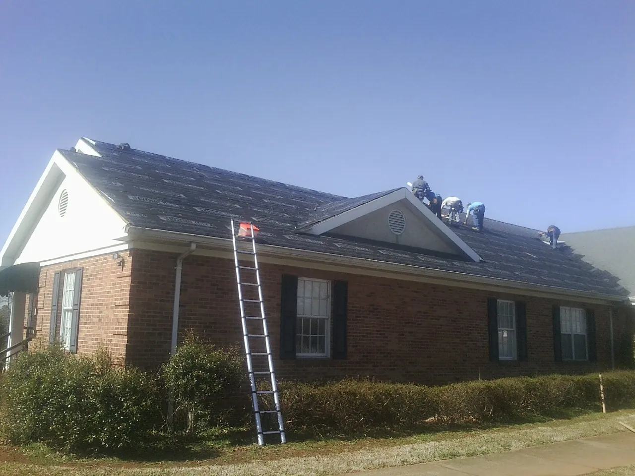 Crew tearing shingles off a brick ranch roof with a ladder set — Emergency Roof Repair work by our crew in Kure Beach